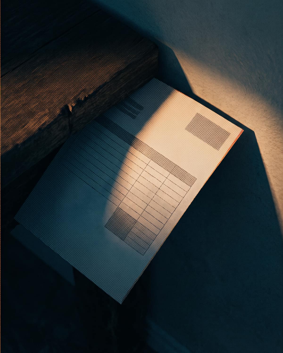 A printed invoice, ruled lines and grey blocks where its details would be, has slipped down between the back of a dark timber desk and the wall. One low shaft of early light catches its top corner; the rest lies in shadow.
